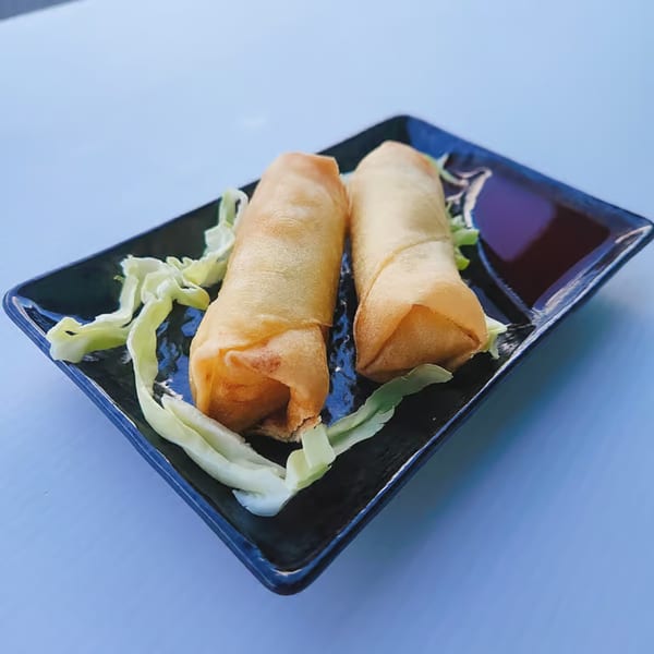 Two golden-brown Chinnie's Kitchen spring rolls with shredded lettuce and dipping sauce on a black plate.
