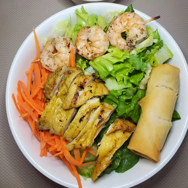 A Chinnie's Kitchen salad with grilled chicken, shrimp skewers, shredded carrots, lettuce, and an egg roll.